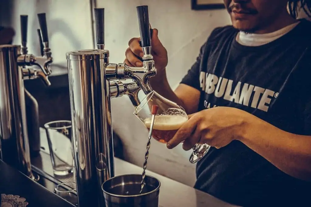 A person fills a tilted glass with beer from a tap at a bar, with foam forming at the top of the drink.