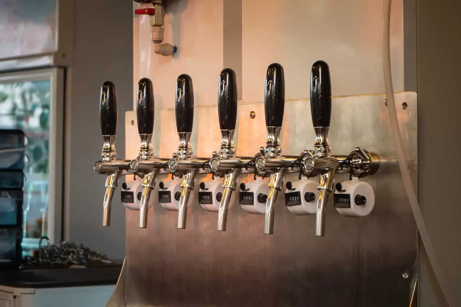 Six beer tap handles are lined up in a row on a stainless steel wall in a bar or brewery setting.