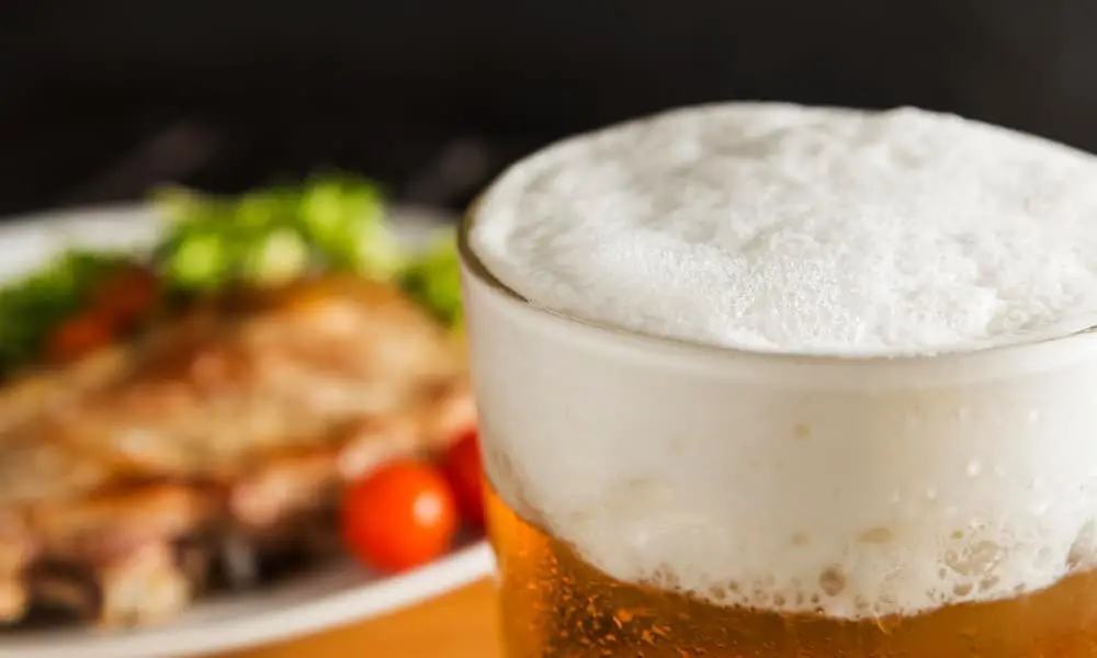 Glass of beer with foamy head and blurred food in background.