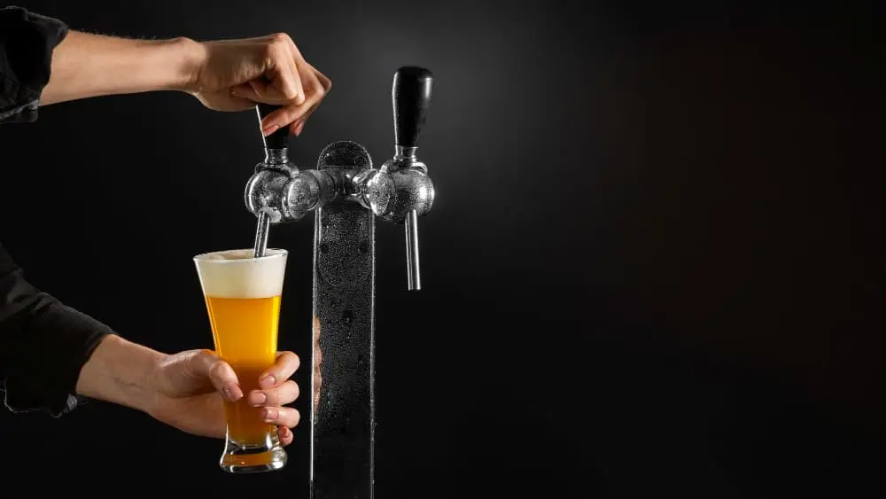 Person pouring draft beer from a tap into a glass, with foam forming on top against a dark background.
