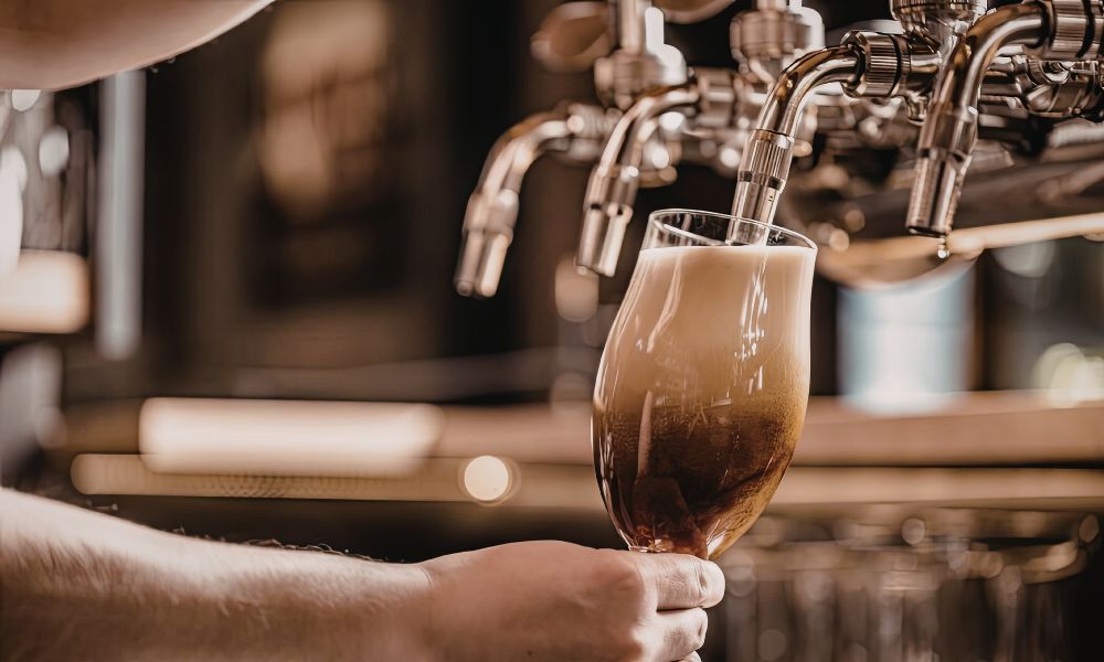 A close-up shot of a person pouring a dark beer with a rich, foamy head from a stainless steel tap